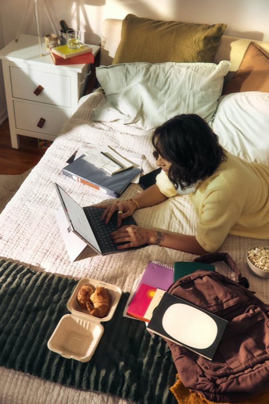 Woman working on a laptop on a bed with books